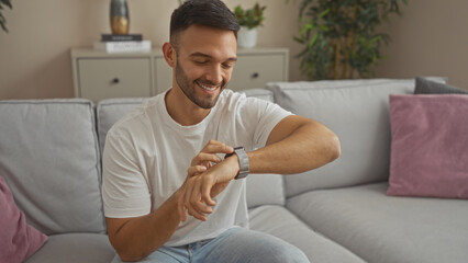 Young hispanic man wearing a white t-shirt and sitting on a gray couch in a living room, smiling while looking at his smartwatch.