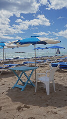 A tranquil beach scene with blue and white umbrellas, sunbeds, a table, and a white chair against a backdrop of the sea and blue sky.
