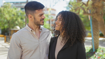 A smiling man and woman embrace lovingly on a sunny day in a city park surrounded by greenery.