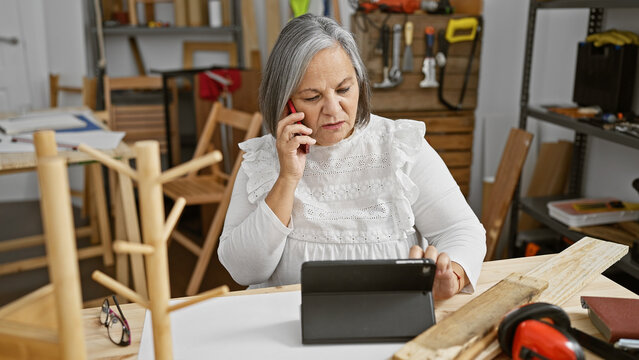 A mature woman speaks on the phone while using a tablet in a well-equipped carpentry workshop