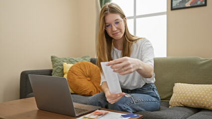 A young adult woman examines a receipt indoors, with a laptop and cozy home decor in the background.