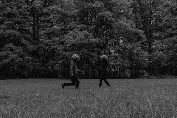 A couple with pumpkin head costumes running towards each other on a green lawn against yellow October forest on a background. Black and white