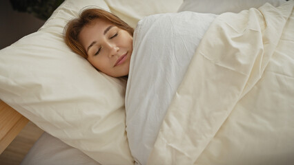 A young caucasian woman sleeps peacefully under a white duvet in a cozy, well-lit bedroom setting.