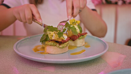 A young woman garnishes eggs benedict in a modern cafe setting, highlighting food, leisure, and pink decor.