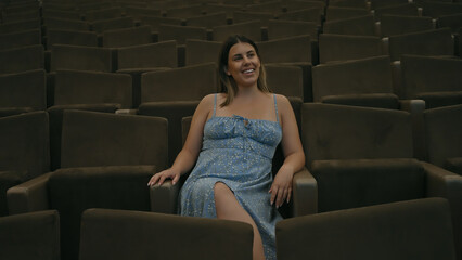 Obraz premium A smiling brunette woman sits alone in a cinema hall wearing a blue dress.