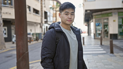 A casual young asian man wearing a jacket stands confidently on an urban city street.