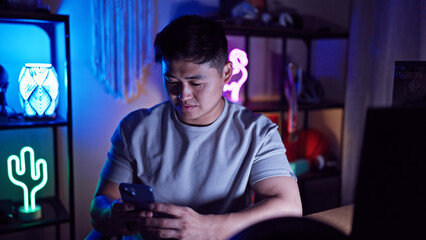 A young asian man checks his smartphone in a neon-lit gaming room at night, depicting technology and lifestyle. © Krakenimages.com
