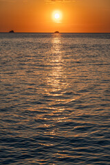 The sun sets over Cala Conta beach in Ibiza, Spain, casting a warm golden glow across the sky. Its reflection sparkles on the textured dark sea