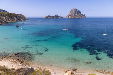 Obraz premium erial view of Cala d Hort beach, Ibiza, highlighting clear emerald waters, sandy beach, anchored sailboats, and a scenic view of Es Vedra and Es Vedranell islands, Spain