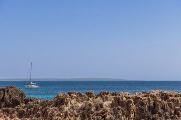 Ibiza rocky coastline with an emerald sea, featuring a distant anchored yacht under a bright blue sky. The scene captures the peaceful and natural beauty of the island