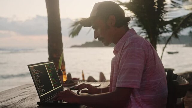 Software developer works on laptop at beach cafe, programming with sea view at sunset. Male coder types code, enjoys remote job with ocean background. Freelancer creates script, relaxed at seaside.