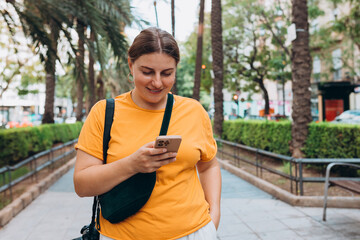 Obraz premium Positive cheerful woman dressed in casual look holding smartphone gadget for communicate on the city street. 30s Woman holding on cell smartphone while relaxing outdoors, Urban lifestyle concept