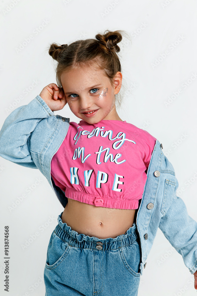 Wall mural Stylish kid girl model with buns wearing jeans and pink t-shirt posing on white studio background