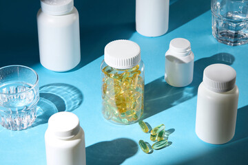Mockup medicine bottles in different sizes evenly arranged on blue table top, with a transparent jar of supplement standing out in center and two water glasses placed on the edges. High angle shot