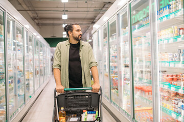 A young man is shopping at the supermarket. A guy walks with a cart between refrigerators with dairy products in a large store