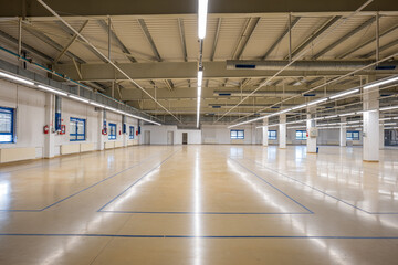 Closed down empty factory interior manufacturing hall with tall ceiling, concrete beams. Wide angle shot, ceiling mounted neon lights, no people