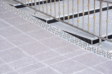 Gray flat tile floor for wheelchair with steps of elevated walkway and stainless steel railing on the ramp area in entryway of outdoors public restroom, perspective side view