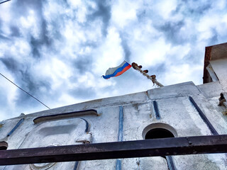 A damaged Russian flag waves atop a ship, highlighting battle scars under a cloudy sky. The Russian...