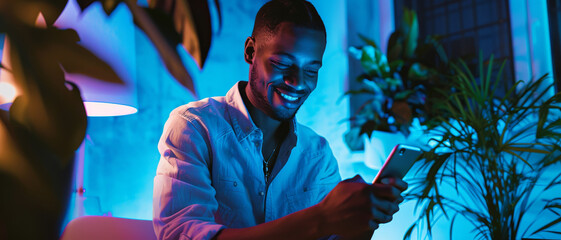 A young man in a cozy, dimly-lit room smiles warmly as he checks his phone, surrounded by house plants and bathed in a serene blue light.