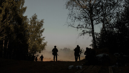 Silhouettes of a group of infantrymen with machine guns in the shade of trees in the forest against...