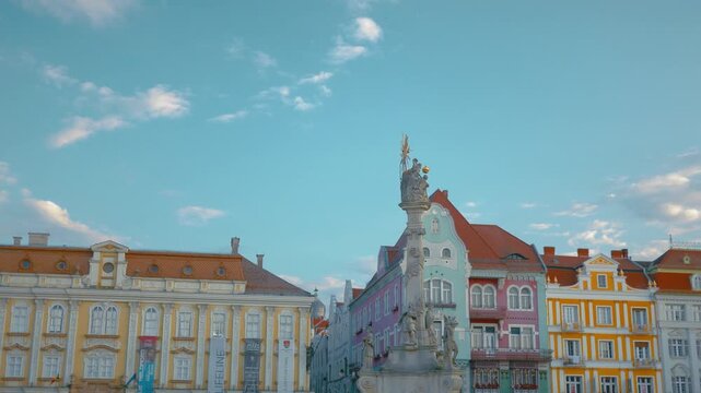 Pan right shot of a column-style monument and pastel colours of old buildings in the background. The location is Union Square in the city centre of Timisoara, Romania.