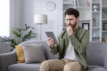 Man seated on sofa reading tablet at home. Thoughtful expression while using digital device. Modern living room background. Concept of technology, leisure, home office, remote work, reading, phone