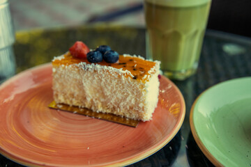 A single slice of coconut cake sits on a pink plate, topped with a layer of passionfruit glaze and two blueberries. A glass of iced tea sits nearby, and the scene is set in a cafe.