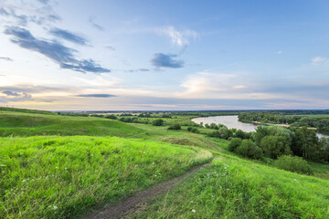 Obraz premium A grassy path meanders through a rolling landscape, leading to a view of a winding river surrounded by trees. The sky is a soft blue with white clouds.