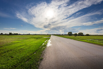 A captivating stretch of road leads through vibrant green fields, embraced by a vast, cloud-dotted sky, capturing the essence of freedom and tranquility.