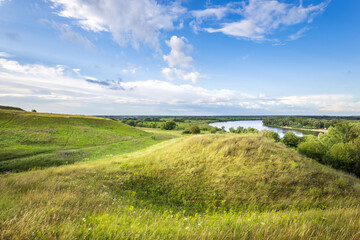 Obraz premium Rolling green hills gently slope toward a calm river, under a bright blue sky scattered with fluffy white clouds, creating a peaceful and natural oasis.