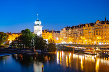 The Sitkov Water Tower stands illuminated on the Masaryk Embankment in Prague, Czechia, as dusk descends. The tower is reflected in the tranquil waters of the Vltava River.