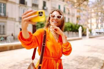 Fototapeta premium Happy attractive young woman posing to camera make selfie smiling at sunlight on street. Travel, tourism, technology, blogging, communication concept.