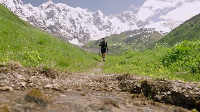 Female tourist with backpack hikes through Jangi-Tau valley in Svaneti. The traveler explores nature with trekking poles and enjoys mountain views. Snow-capped peaks in background. Slow motion.