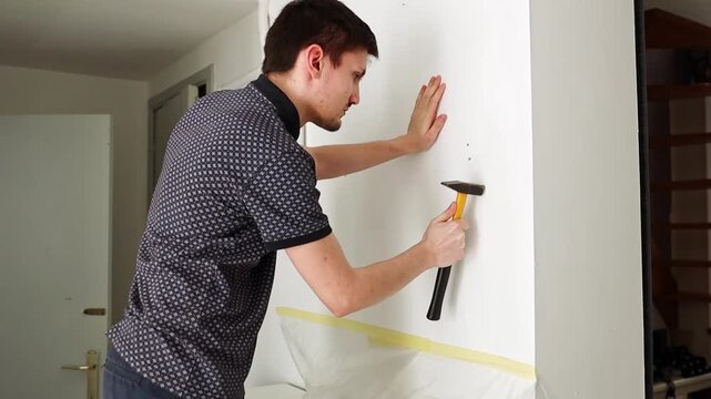 A young man hammers a dowel into a hole on the wall.