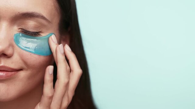 A close-up image of a serene woman applying blue hydrogel under eye patches for skincare on a clear background.