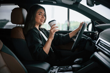 White cup of fresh drink in hand. Beautiful businesswoman is in the new car