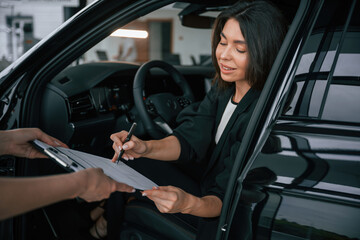 Writing by a pen. Process of buying new car, woman is signing documents while sitting in a new...