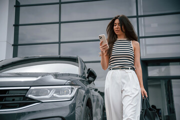 With smartphone. Attractive young woman is outdoors near her brand new car