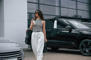 Walking holding drink. Attractive young woman is outdoors near her brand new car