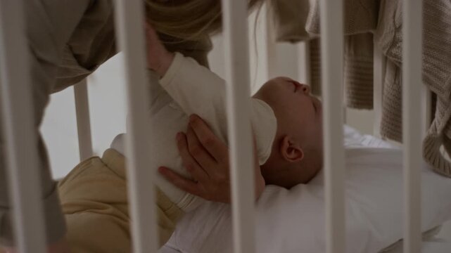 Medium shot of unrecognizable young mother softly and securely placing baby into crib leaving little child safely sleeping on white pillow