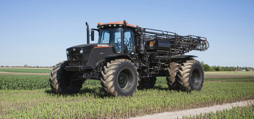 Obraz premium Large Agricultural Sprayer Tractor Working In Green Crop Field Under Clear Blue Sky