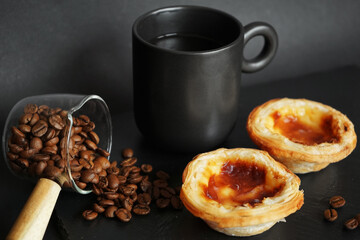 Egg tarts next to a black cup of coffee and scattered coffee beans on a dark background