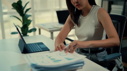 Stressed businesswoman reviews financial reports at office desk with laptop. Analyzing documents in professional setting during daytime