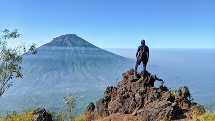 a man standing on a rock looking at a mountain, mount sindoro, indonesia