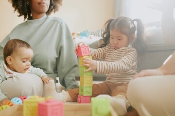 Asian girl trying to built brick tower while baby boy touching her snow white sock, their mothers sitting next to them