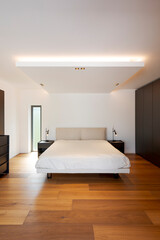 Interior of Bedroom with double bed, bedside tables with lights above. Wooden floor, white walls, no one inside.