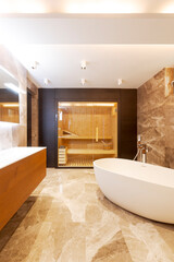 Modern bathroom with marble walls and floor. Bathtub in the middle of the room. In the background you can see a sauna with glass door.