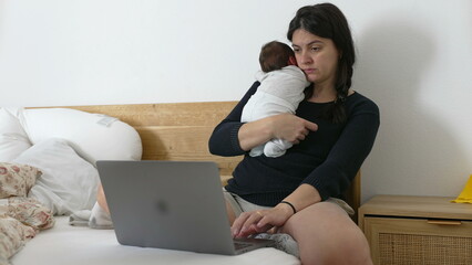 Focused mother multitasking by holding newborn baby and working on a laptop, challenges of...