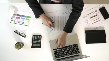 A man is sitting at a desk with a green tablet in front of him. The tablet is on a white surface with a laptop and a calculator nearby. There are also several papers on the desk
