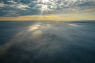 A stunning aerial view of a dramatic cloudscape with the sun's rays breaking through the clouds, casting a serene glow over the scene. The interplay of light and shadow creates a captivating and tranq